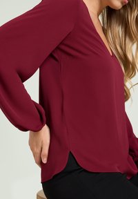 Bordowa bluzka z dekoltem w serek, długimi rękawami i bocznymi rozcięciami, wykonana z lekkiego materiału. Gładka tekstura, delikatnie opada na sylwetkę.