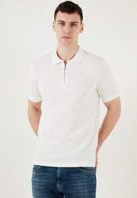 Vit kortärmad polo med dragkedja vid kragen, med en mjuk textur och kontrasterande kragskärning, kombinerad med blå denimjeans.