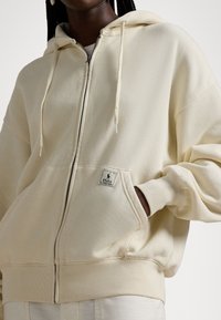 Lekka kremowa bluza z kapturem na zamek, o luźnym kroju, z prążkowanymi mankietami i przednimi kieszeniami. Z przodu małą naszywkę z logo. Miękka w dotyku.
