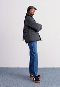 Doudoune gris foncé, boutonnée, portée par-dessus un haut noir. Jean droit bleu et baskets noires avec des semelles en gomme. Casquette grise.