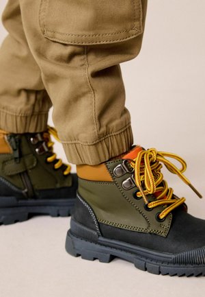 Kind trägt grüne und schwarze Wanderschuhe mit gelben Schnürsenkeln und beige Cargohosen, an den Knöcheln hochgekrempelt, vor neutralem Hintergrund.