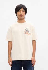 Κρεμ κομπλέ μπλουζάκι από βαμβάκι με κοντά μανίκια, που διαθέτει εκτυπωμένο λογότυπο και σχέδιο στην άνω αριστερή πλευρά του θώρακα. Χαλαρή εφαρμογή και απαλό υφή.