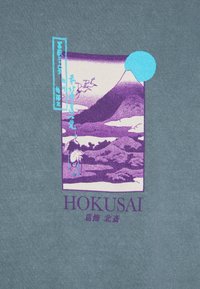 Der graue Stoff weist einen lila und blauen Druck einer Landschaft mit einem Berg, Wasser und einem Kranich auf, neben dem Text "HOKUSAI" in Lila.