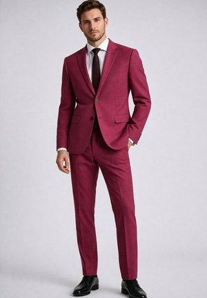 Homme debout portant un costume rouge foncé sur mesure, une chemise blanche, une cravate noire et des chaussures habillées noires sur un fond clair uni.