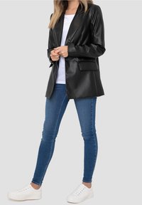Blazer nero in ecopelle con un design classico dei revers, chiusura con un solo bottone e tasche laterali, abbinato a jeans blu e sneakers bianche.