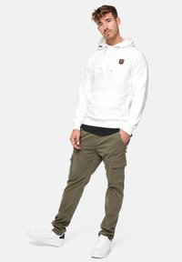 Sudadera blanca con un bolsillo tipo canguro y un parche con el logo marrón, combinada con pantalones cargo verdes oliva que tienen bolsillos laterales y zapatillas blancas.