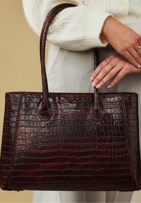 Bruine leren handtas met krokodillenstructuur, dubbele handvatten en een gestructureerd ontwerp. Voorzien van een logo aan de voorkant.
