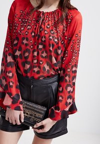 Blusa rossa trasparente con stampa leopardata nera, scollatura arricciata e maniche lunghe. Abbinata a pantaloni corti di pelle nera e una clutch nera con borchie.