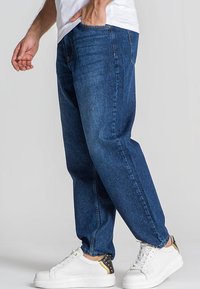 Jeans de color azul oscuro en un corte relajado, con un clásico diseño de cinco bolsillos y un sutil desgaste. Combinados con zapatillas blancas y una camiseta blanca sencilla.