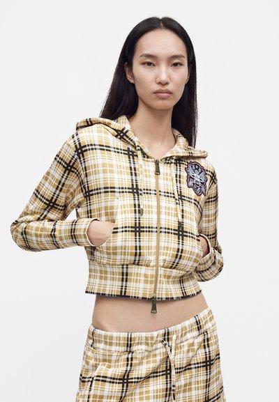 Νεαρή γυναίκα φοράει cropped hoodie με κίτρινο και μαύρο καρό, με τα χέρια στις τσέπες και ταιριαστό καρό παντελόνι σε απλό φόντο.