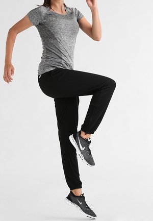 Person trägt graues T-Shirt, schwarze Jogginghose und schwarze Nike-Sneaker und hebt ein Knie in der Luft vor weißem, einfachem Hintergrund.