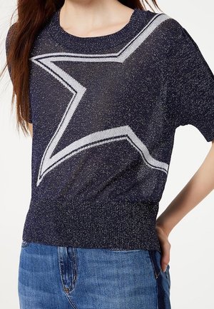 Kobieta nosząca granatowy, krótki sweter dzianinowy z dużym białym wzorem gwiazdy, w połączeniu z niebieskimi jeansami denim.