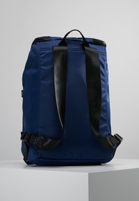 Ciemnoniebieski plecak z czarnymi, regulowanymi paskami na ramiona i uchwytem do przenoszenia na górze, stojący pionowo na białym piedestale na szarym tle.