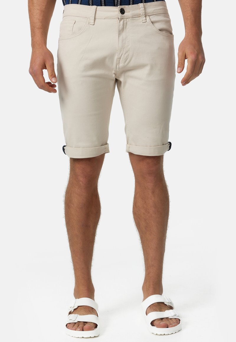 Modèle masculin portant un short beige retroussé, une chemise rayée marine et des sandales blanches à double bride, se tenant devant un fond uni.