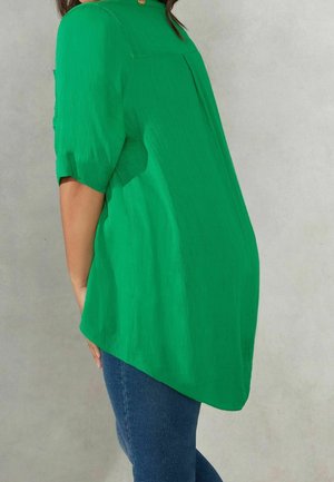 Groene, lichte blouse met een ontspannen pasvorm, korte mouwen en een hoge-lage zoom. Bevat een achterhempièce en subtiele textuur. Gedragen met blauwe jeans.