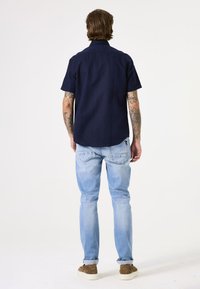 Marineblauw kortarmshirt, ontspannen pasvorm, gemengde stof, gecombineerd met lichtblauwe jeans, opgerolde mouwen, tan sneakers, zichtbare tattoos op de armen.