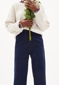 Maglione bianco a coste indossato con jeans a gamba dritta blu scuro, mentre tiene steli verdi legati con spago, adornati con braccialetti di perline colorate.