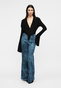 Nő áll, egyik kezét a csípőjén tartja, fekete mély V-nyakú fodros felsőt és bővülő, batikolt kék farmert visel.