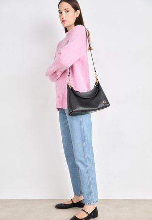 Black leather shoulder bag with curved shape, gold hardware accents, and a logo detail. Model wears a pink fuzzy sweater and blue jeans.