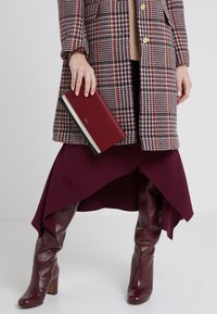 Kvinde iført burgunder læderstøvler, asymmetrisk burgunder nederdel, ternet frakke, holder en burgunder clutch med hvide og sorte detaljer.