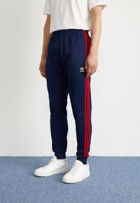 Marina atletiska byxor med röda sidostraper, elastisk midja och en guldig adidas-logotyp. Kombinerat med vita sneakers på en blå texturerad yta.