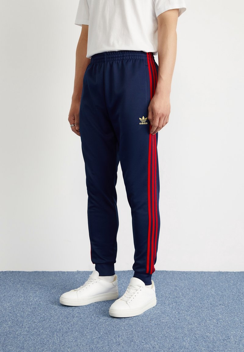 Marina atletiska byxor med röda sidostraper, elastisk midja och en guldig adidas-logotyp. Kombinerat med vita sneakers på en blå texturerad yta.