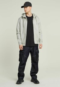 Haut à capuche zippé gris clair avec cordons de serrage sur un t-shirt noir, associé à un pantalon cargo bleu foncé comportant plusieurs poches et des coutures contrastantes.