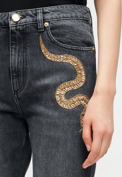 Zwarte spijkerbroek met gouden kralenslangversiering die langs het bovenbeen kronkelt, gedragen door een persoon met een ontspannen hand langs het lichaam.