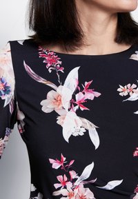 Nainen, joka on pukeutunut mustaan mekkoon, jossa on vaaleanpunaisia, valkoisia ja purppuranvärisiä kukkakuvioita, lyhyet tummat hiukset ja hieman käännös päässä, joka paljastaa kaulan ja leuan.