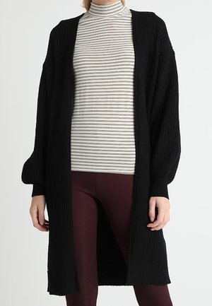 Femme portant un long cardigan noir sur un col roulé rayé blanc et gris et un pantalon texturé bordeaux.