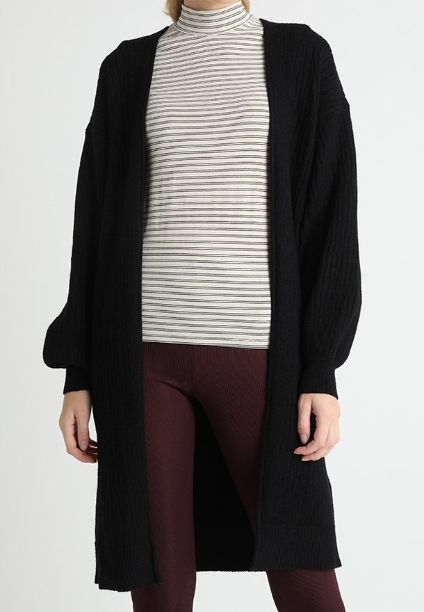 Femme portant un long cardigan noir sur un col roulé rayé blanc et gris et un pantalon texturé bordeaux.