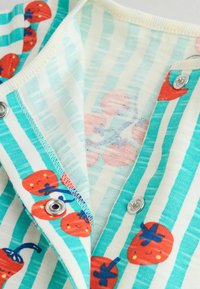 Detailný záber na pruhované detské oblečenie s úsmevnými červenými potlačami paradajok a kovovými cvokmi pozdĺž otvorenia.