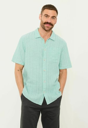 Mann mit Schnurrbart, der ein hellgrünes Kurzarmhemd mit Knöpfen und schwarze Hose trägt, steht mit den Händen in den Taschen vor weißem Hintergrund.