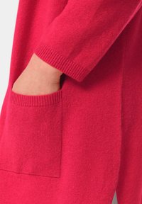 Un pull en tricot rose vif avec une poche latérale, présentant des poignets côtelés et une texture douce. Montre un gros plan du tissu et des détails de la poche.
