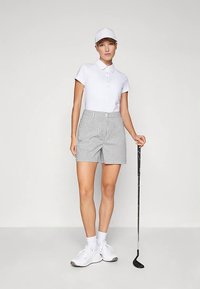 Balta polo krekls ar pogām, pelēkas un baltas rūtainas šorti, balta cepure, balti sporta apavi, turot golfa nūju.