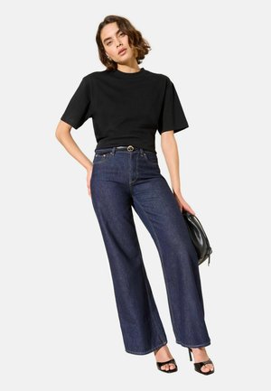 Musta lyhythihainen t-paita yhdistettynä korkeavyötäröisiin tummansinisiin leveälahkeisiin farkkuihin. Mustat korkokengät ja musta käsilaukku täydentävät asun.