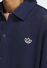 Marinblå kortärmad polo tillverkad av strukturerad meshtyg, med en knappad krage och en vit broderad logotyp på vänster bröst.