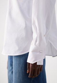 White button-up shirt with a smooth texture, featuring a classic collar, long sleeves, and a silver accent on the cuff. Worn with blue jeans.