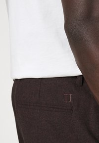 Pantalones marrón oscuro con textura y un discreto detalle bordado en rosa "II", combinados con una camiseta blanca de cuello redondo.