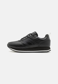 Zapatillas deportivas negras con un acabado de cuero suave, suela blanca y suela de goma negra texturizada; cuentan con cordones y una pequeña etiqueta en el talón.