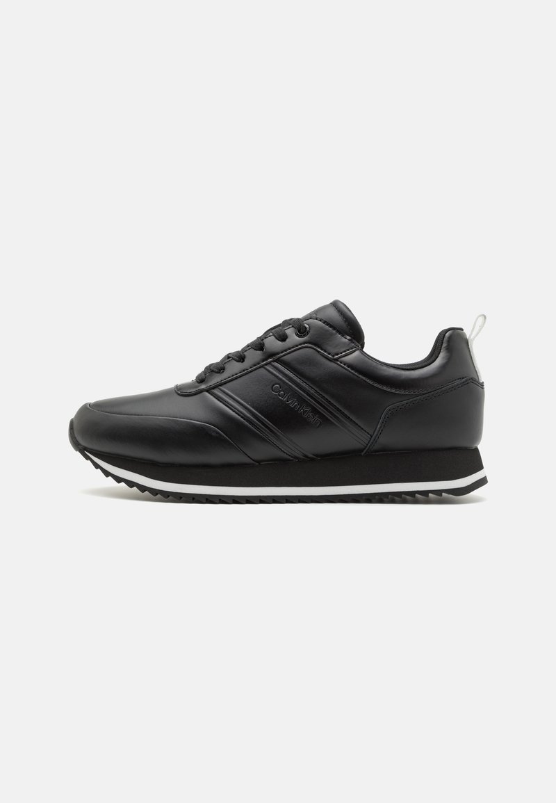 Zapatillas deportivas negras con un acabado de cuero suave, suela blanca y suela de goma negra texturizada; cuentan con cordones y una pequeña etiqueta en el talón.