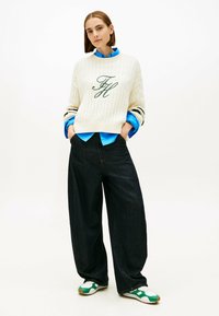 Pulover crem cu tricotaj cablu și inițiale verzi brodate, asortat cu o cămașă butonată de un albastru deschis, blugi negri cu picioare largi și pantofi sport cu accente verzi.