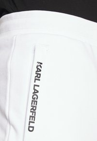 Bílá sportovní tkanina s elastickým páskem, na bočním švu svislá černá nášivka s nápisem "KARL LAGERFELD".