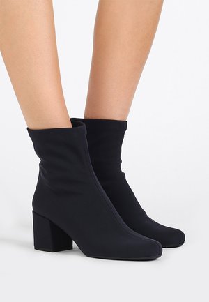 Bottes chaussettes noires à mi-mollet avec talons blocs, portées sur des jambes nues, sur un fond blanc uni.