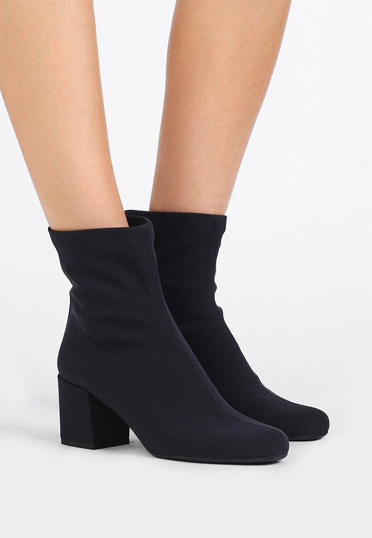 Bottes chaussettes noires à mi-mollet avec talons blocs, portées sur des jambes nues, sur un fond blanc uni.