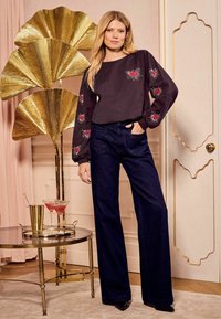 Črna dolga majica s dolgimi rokavi z vezenim rdečim srčkom, v kombinaciji z temno modrimi širokimi hlačami. V ozadju zlate dekorativne ročne lepe in set za koktajle.