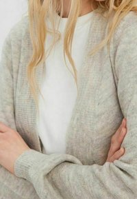 Jasnoszary kardigan o prążkowanej fakturze, otwarty z przodu, z długimi rękawami. Nosi się go na białą bluzkę, o luźnym kroju i miękkim materiale.