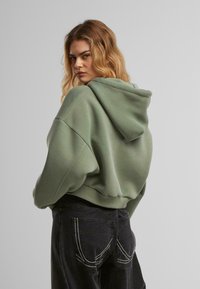 Hanorac scurt verde cu glugă reglabilă, textură moale și croială oversize, asortat cu blugi închiși la culoare, având cusături contrastante pe buzunare.