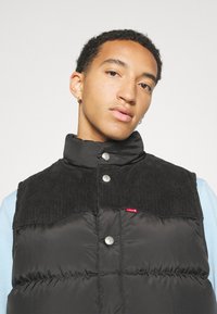 Crni puffer prsluk s dijelom od kordura na ramenima, dugmadi na kopčanje i malim crvenim Levi's logotipom. Ima visoki ovratnik i glatku teksturu.