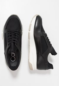 Baskets de sport noires avec un dessus lisse, un bout arrondi et une semelle blanche épaisse. Les lacets présentent un design standard et sont placés au centre.